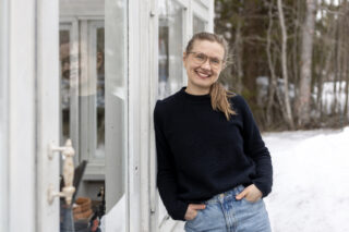 noin 30-vuotias hymyilevä nainen, Riikka, vaaleanruskeat hiukset, tumman sininen neule, nojaa talon kuistiin. Takana puunrunkoja.