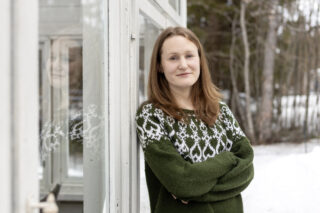 Noin 30-vuotias nainen, punertavat pitkät hiukset, vihreä neulu, katsoo pienesti hymyillen kameraan. Liisa nojaa valkoiseen kuistiin, takana puun runkoja.